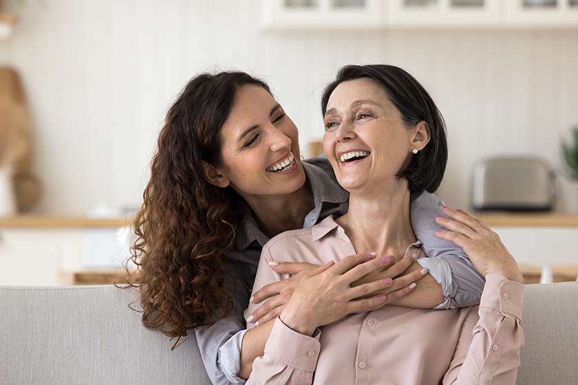 Happy Mothers Day celebration, family ties and unconditional love. Cheerful beautiful grown up millennial daughter cuddling from behind her lovely mature mother, express care, support and affection 